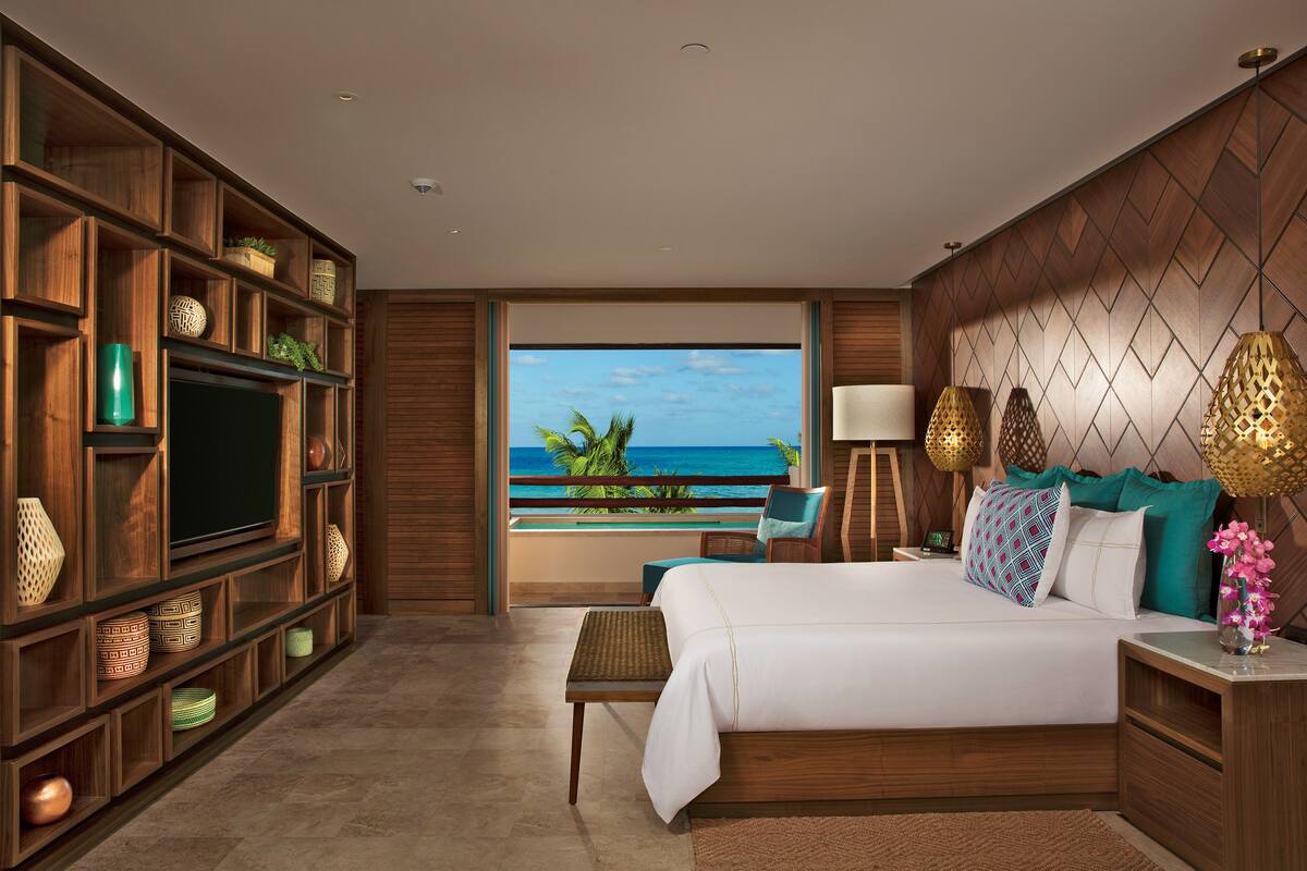 A hotel room with a large bed, a television, wooden shelving with decorative items, a balcony with a view of the ocean, and a nightstand with lamps.