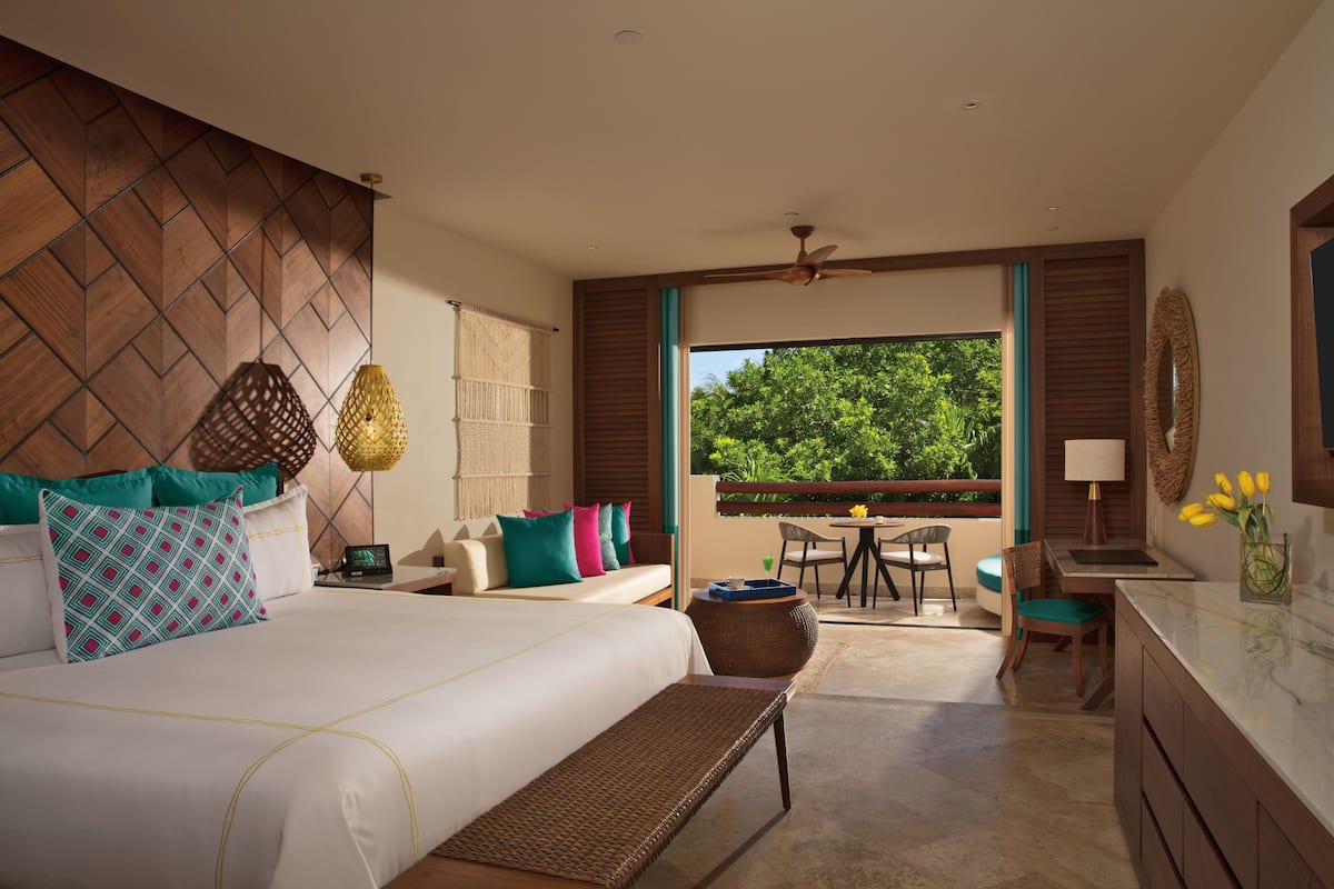 A modern hotel room with a large bed, a balcony with a table and chairs, and a view of greenery outside.