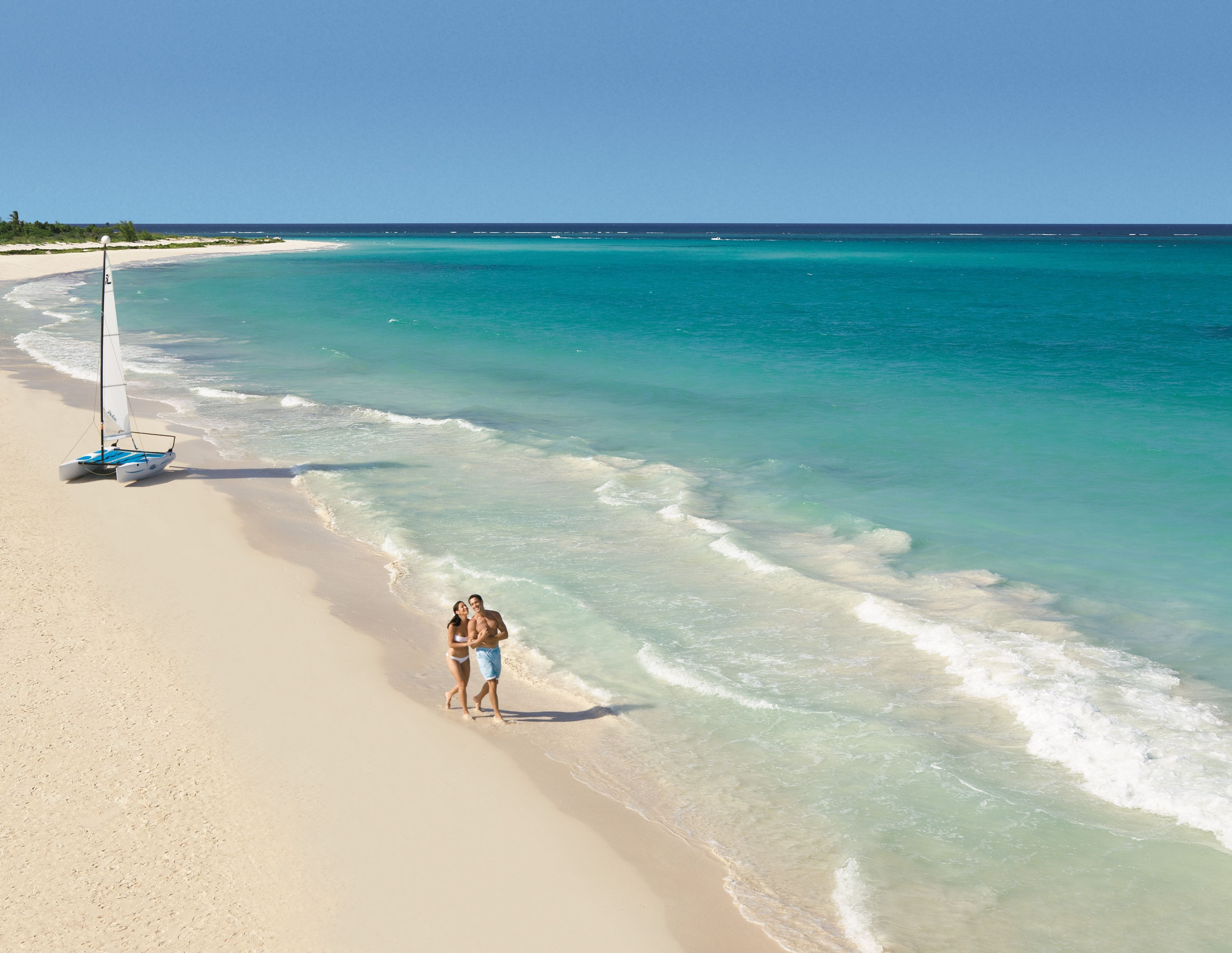 A beach with clear turquoise water, a sailboat, and a couple walking along the shore.