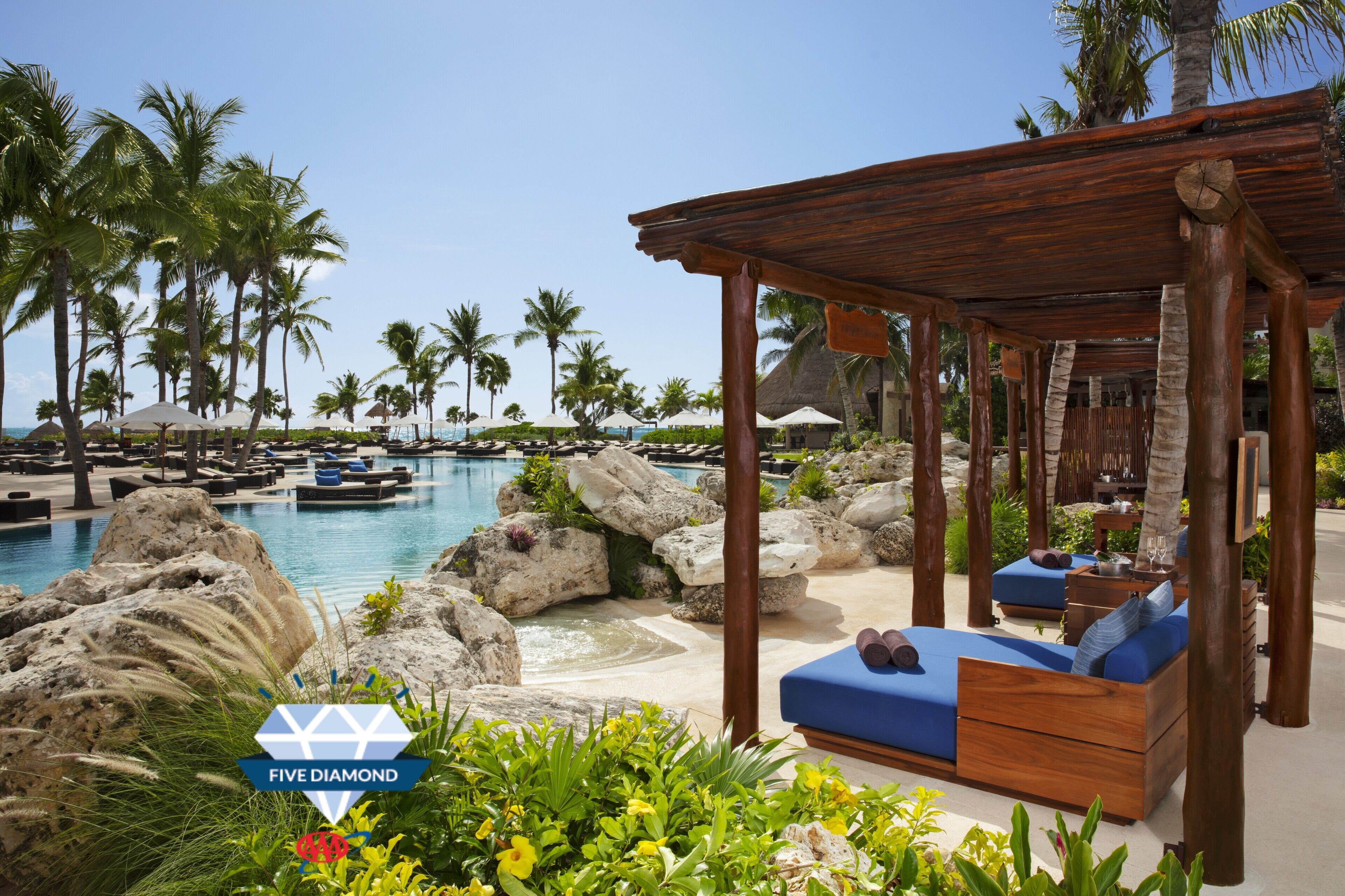 A resort pool area with wooden cabanas, lounge chairs, and palm trees.