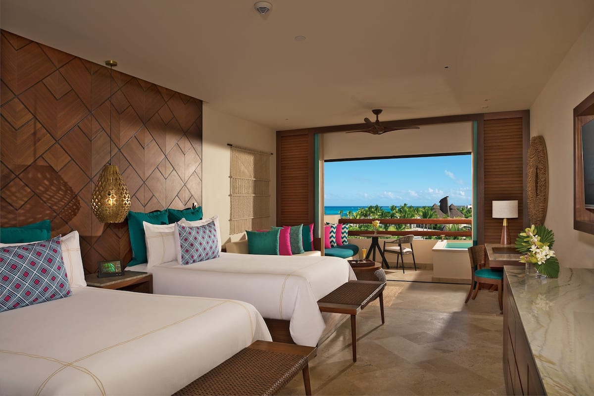 A hotel room with two beds, a balcony with a view of the ocean, a dining area, and a bathroom.