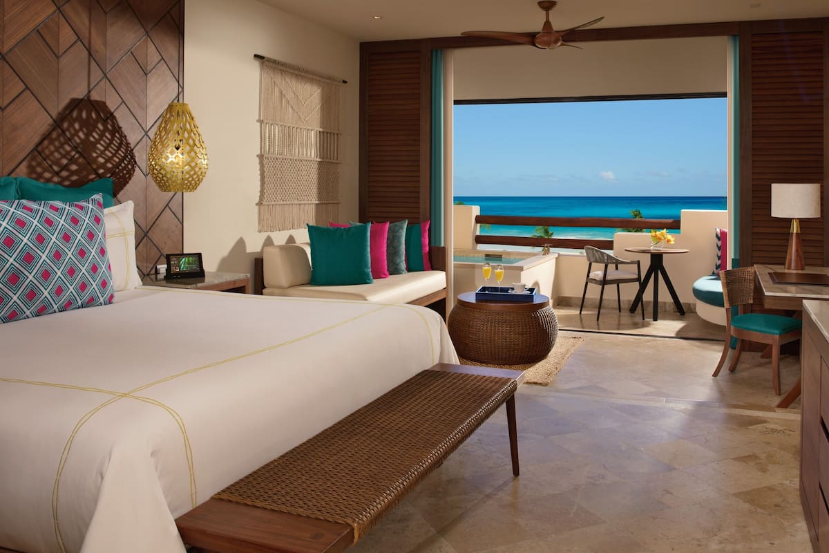 A hotel room with a large bed, a desk, a chair, a small table, and a view of the ocean.