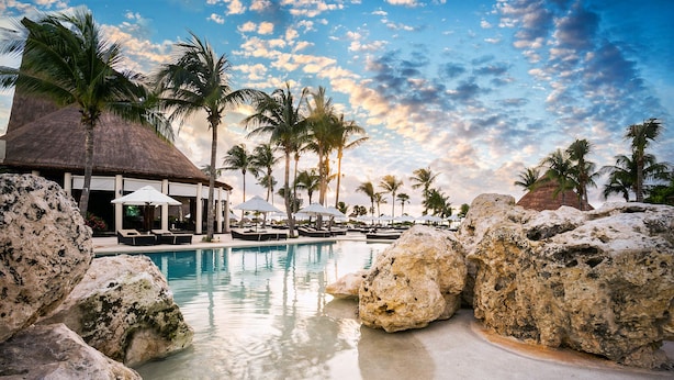 A resort pool area with large rocks, palm trees, and thatched-roof structures. - Secrets Maroma Beach Riviera Cancun - Adults Only - All inclusive (Playa del Carmen)
