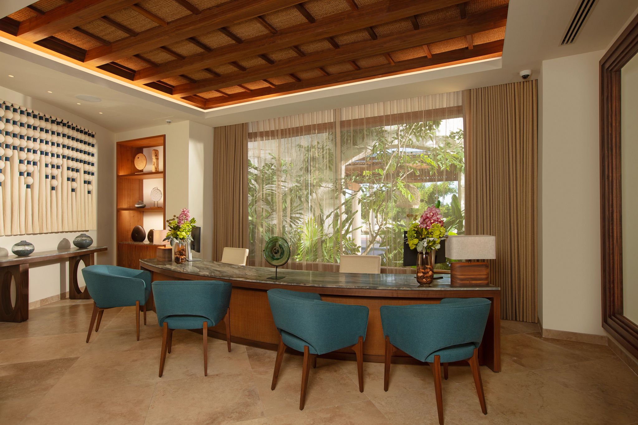 A modern kitchen with a wooden ceiling, a bar area with blue chairs, and a view of greenery outside.