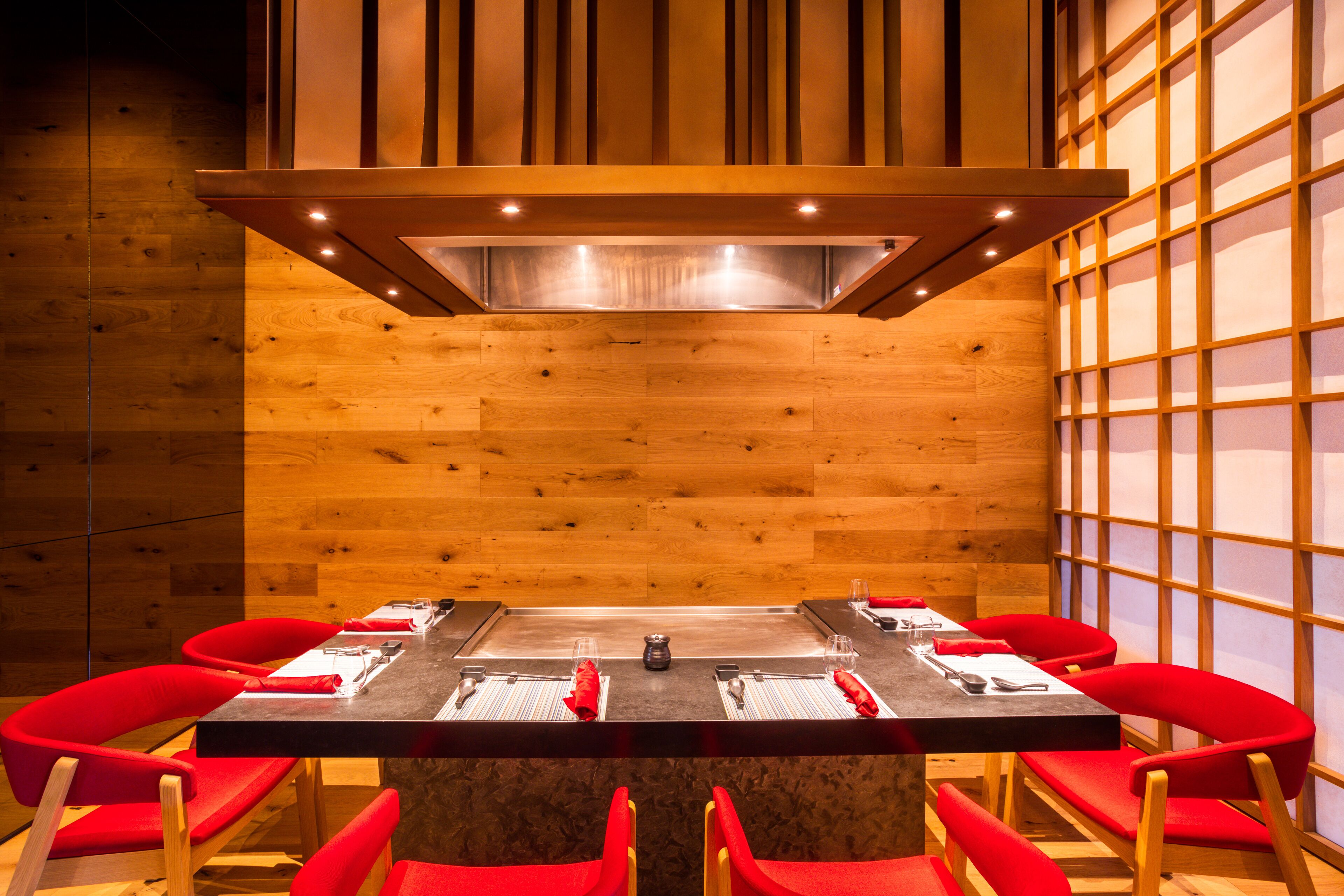 A dining area with a long table set for six, surrounded by red chairs, in a room with wooden walls and a modern design.