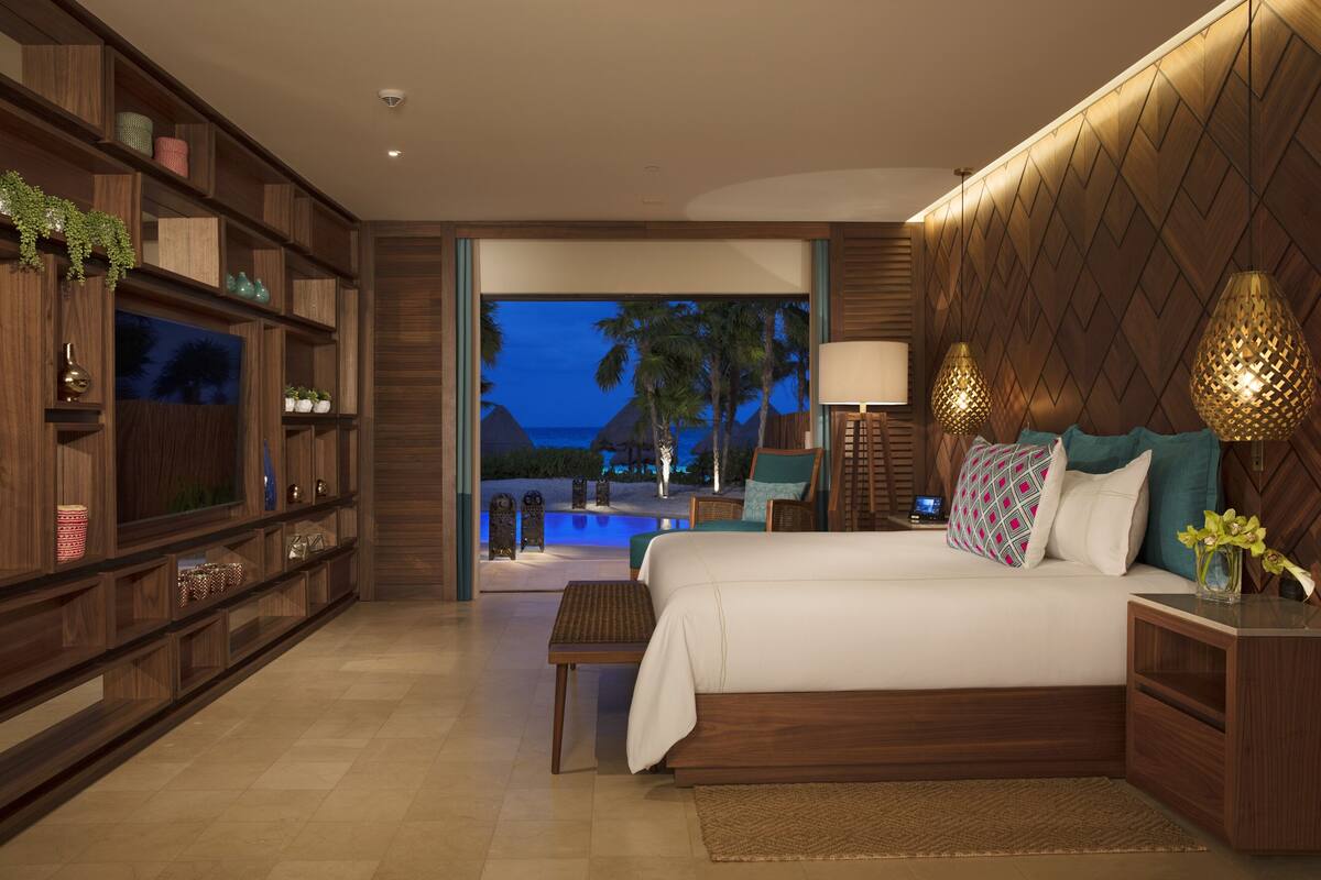 A modern hotel room with a large bed, wooden shelving, a television, and a view of a pool and palm trees at dusk.