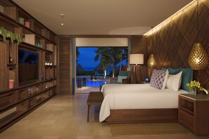 A modern hotel room with a large bed, wooden shelving, a television, and a view of a pool and palm trees at dusk.