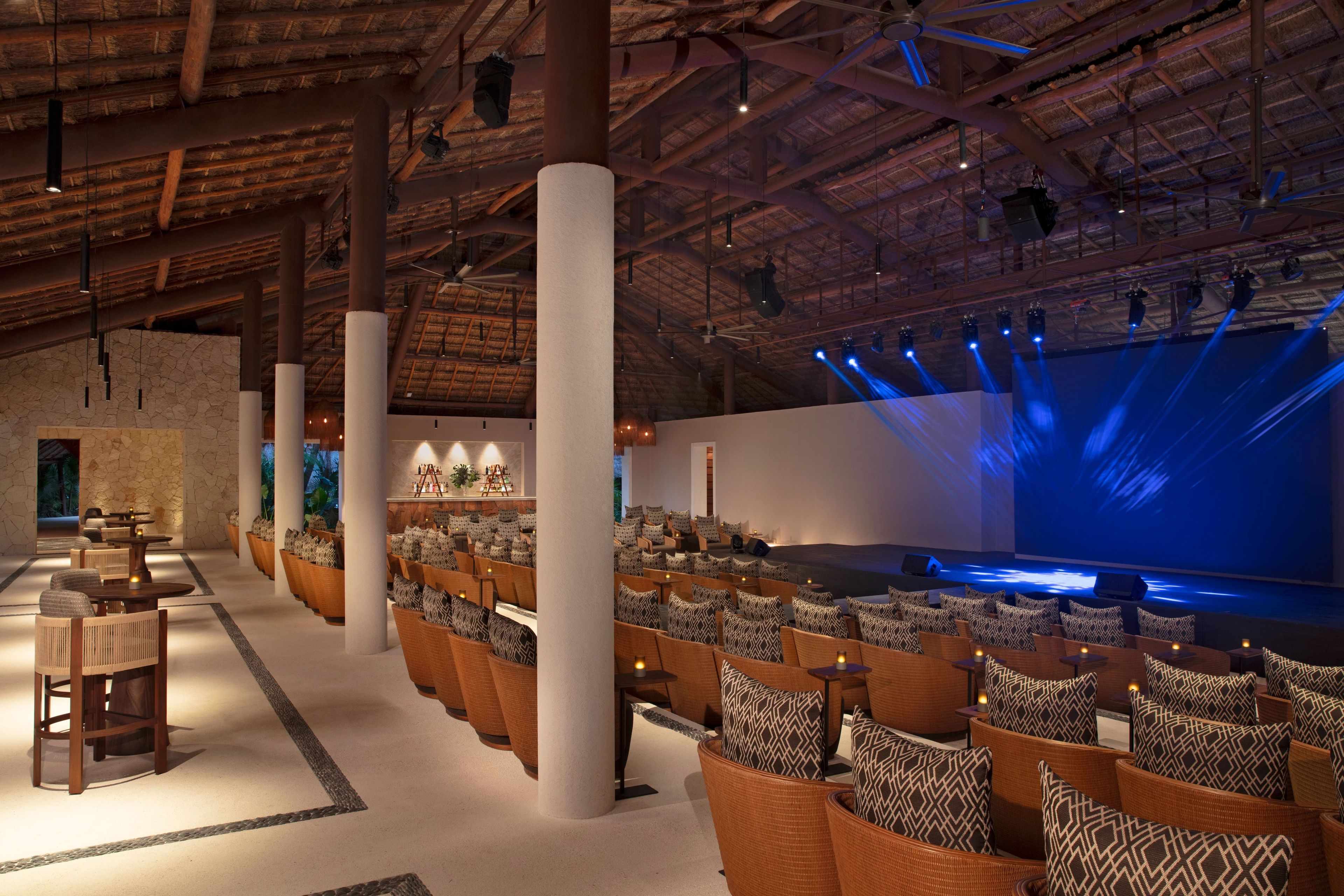 A spacious indoor venue with high ceilings, a stage with blue lighting, and rows of seating with patterned cushions.