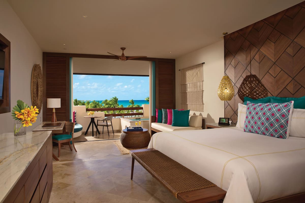 A modern hotel room with a large bed, a desk, a chair, and a balcony with a view of the ocean.