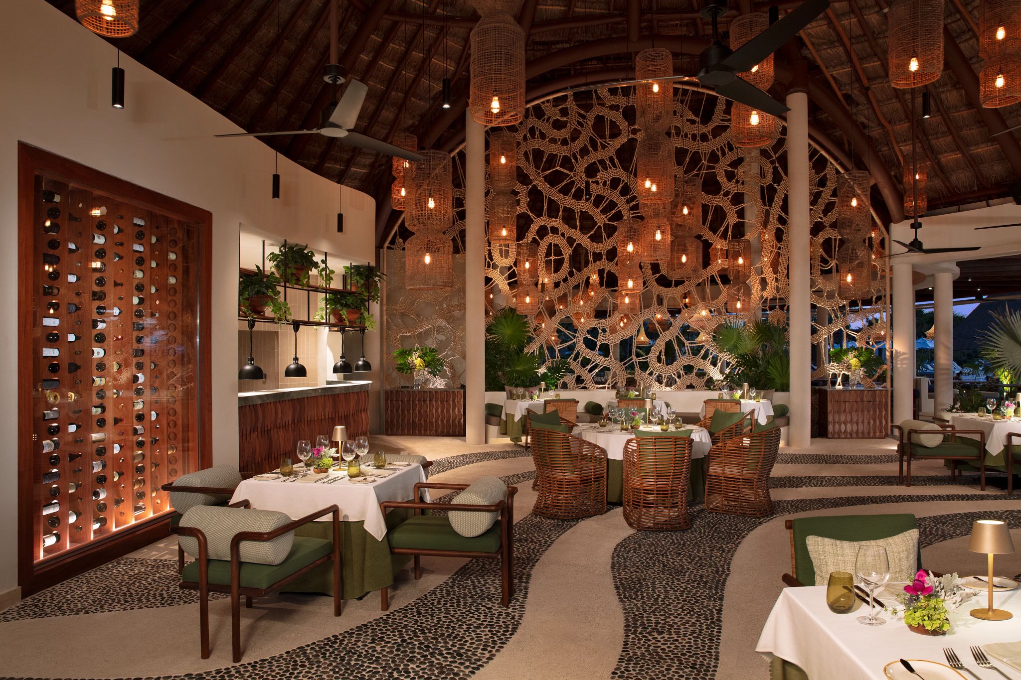 A restaurant with a thatched roof, wicker furniture, and a wall of wine bottles.
