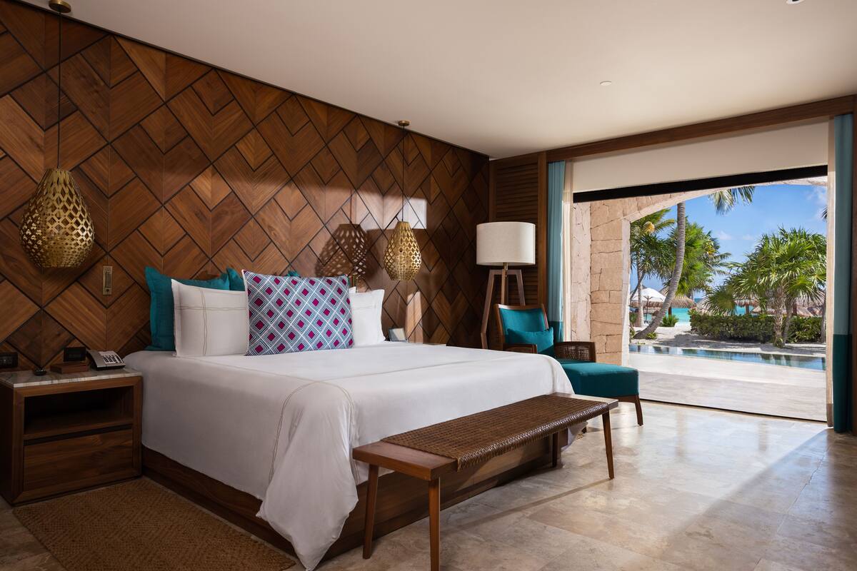 A hotel room with a wooden herringbone wall, a large bed with patterned pillows, a bench, a nightstand with a phone, and a view of a pool and palm trees through a glass door.