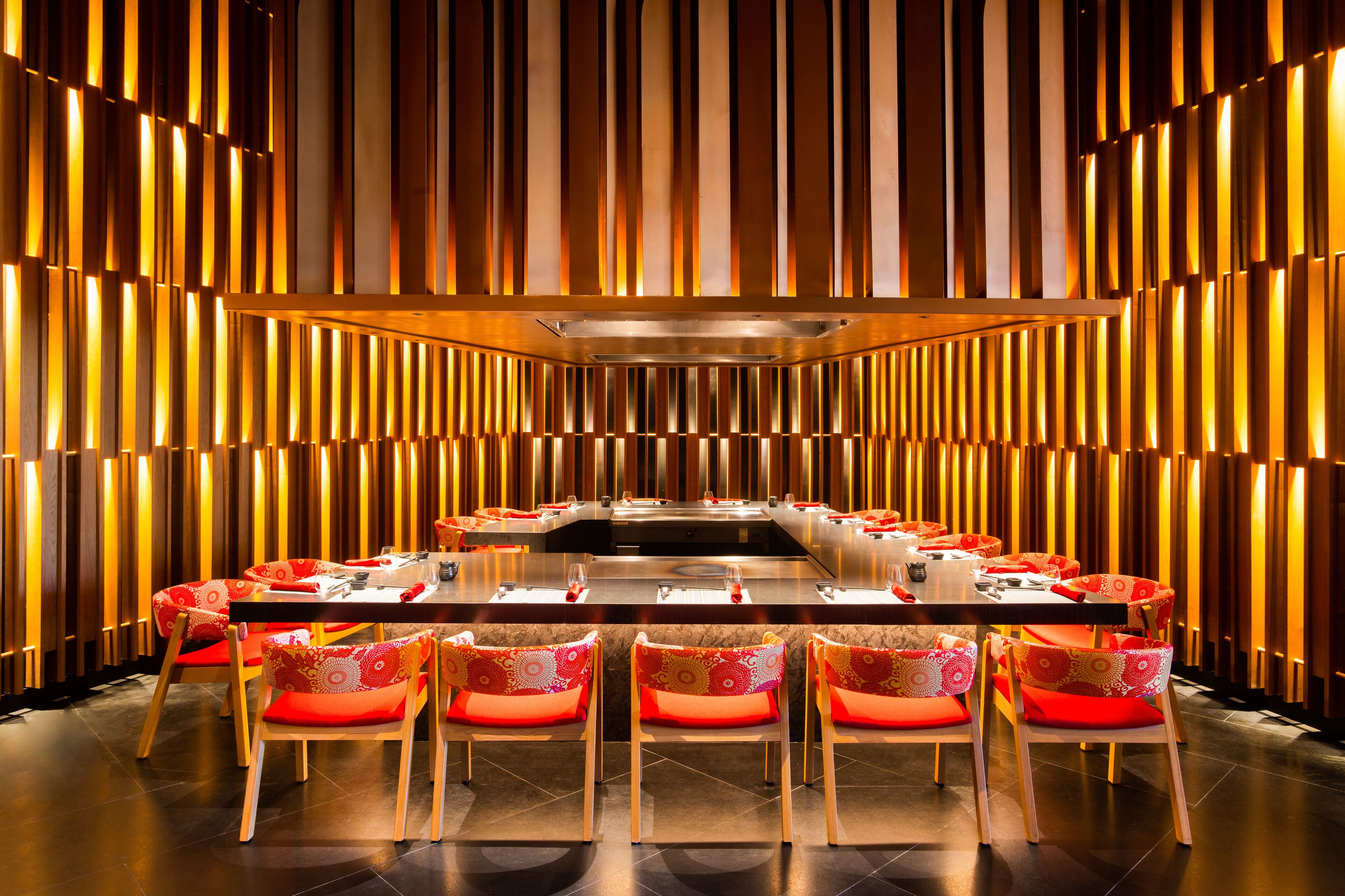A modern dining area with a long table and red upholstered chairs, set against a backdrop of vertical wooden slats.