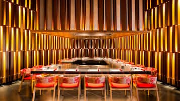 A modern dining area with a long table and red upholstered chairs, set against a backdrop of vertical wooden slats.