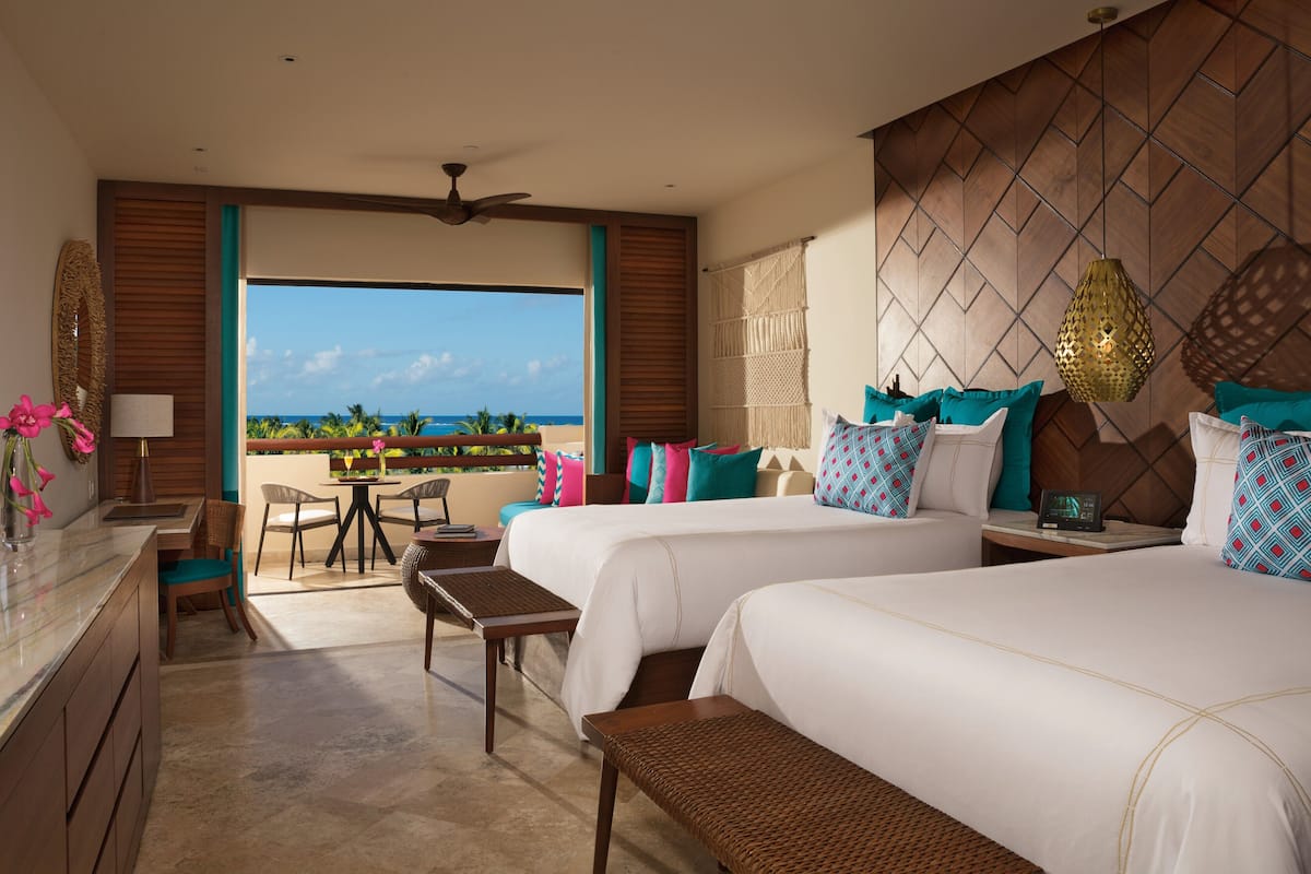 A hotel room with two beds, a desk, a chair, a small table, and a view of the ocean.