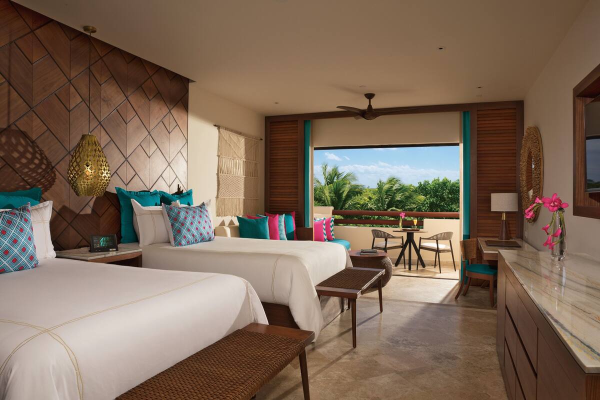 A hotel room with two beds, a balcony with a table and chairs, and a view of palm trees.