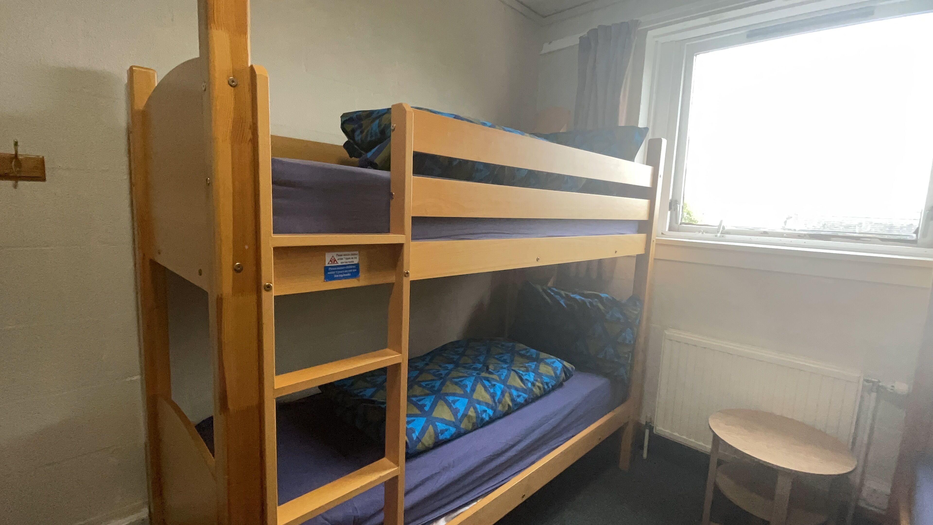 Una cama litera con una cama inferior y una superior, ambas con ropa de cama de estampado azul. Junto a la ventana hay una mesita pequeña.