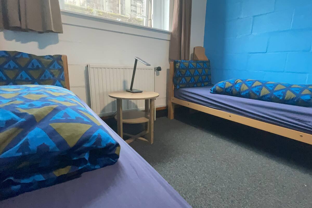 A dormitory room with two beds, a small table, and a window.