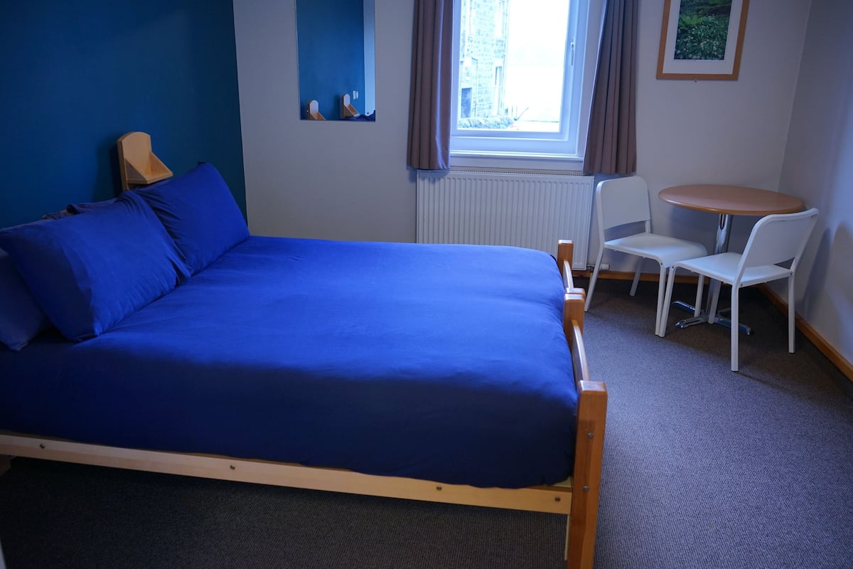 A room with a bed, a small table with two chairs, a window with curtains, and a radiator.