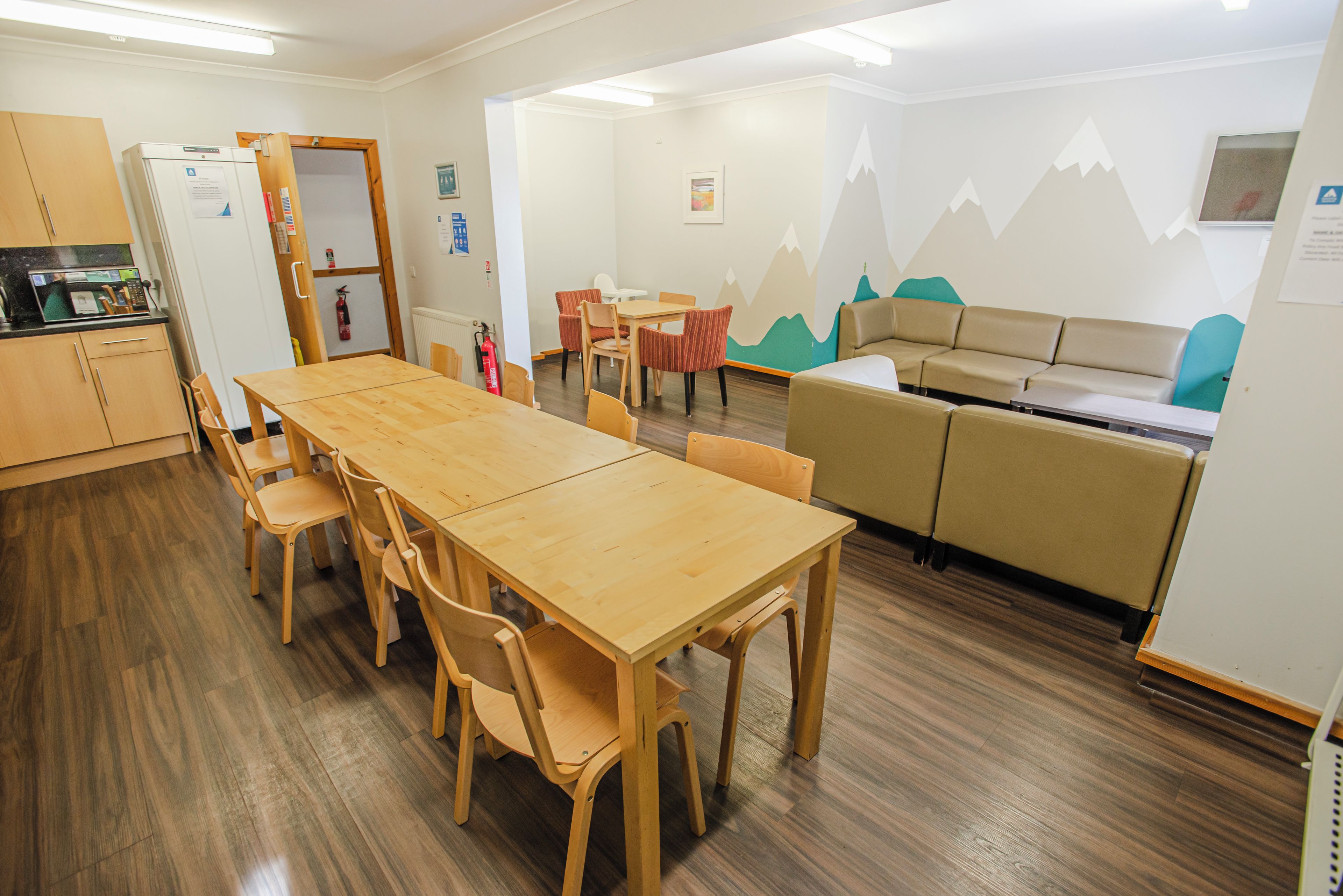Un comedor con mesa y sillas de madera, una kitchenette, un sofá y una pared con un diseño de montaña.