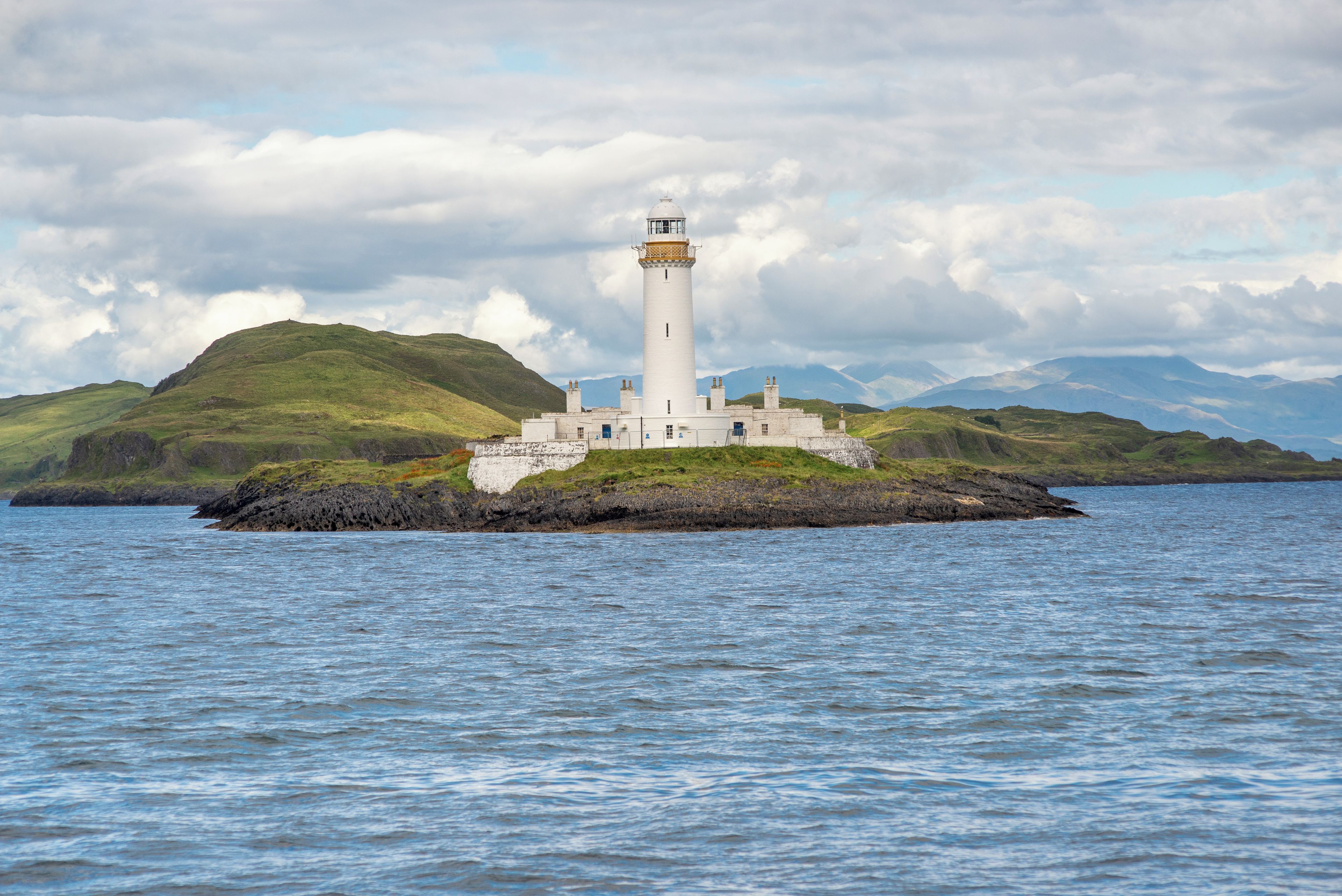 A lighthouse on a small island with a rocky shoreline and a mountainous backdrop.