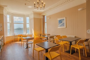 A dining area with multiple tables and chairs, large windows with a view of the sea, and a chandelier.
