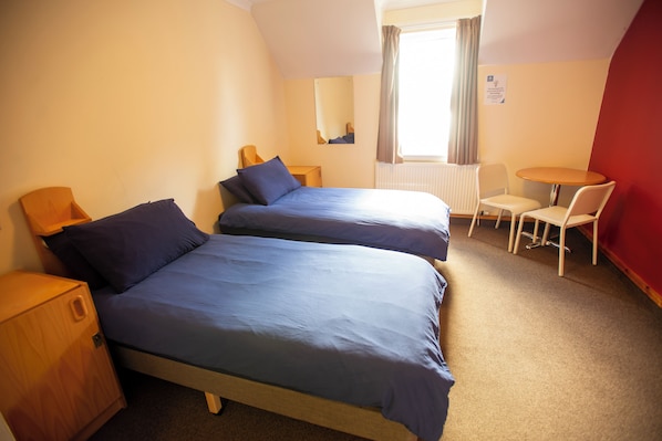 A hotel room with two beds, a small table, a chair, a radiator, and a window with curtains.