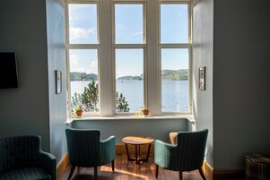 Habitación con un ventanal grande con vistas a un cuerpo de agua y colinas. Hay dos sillas y una mesita en la habitación.