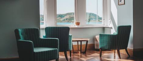 A room with two green upholstered chairs, a small round table, and a view of a coastal landscape through a window.