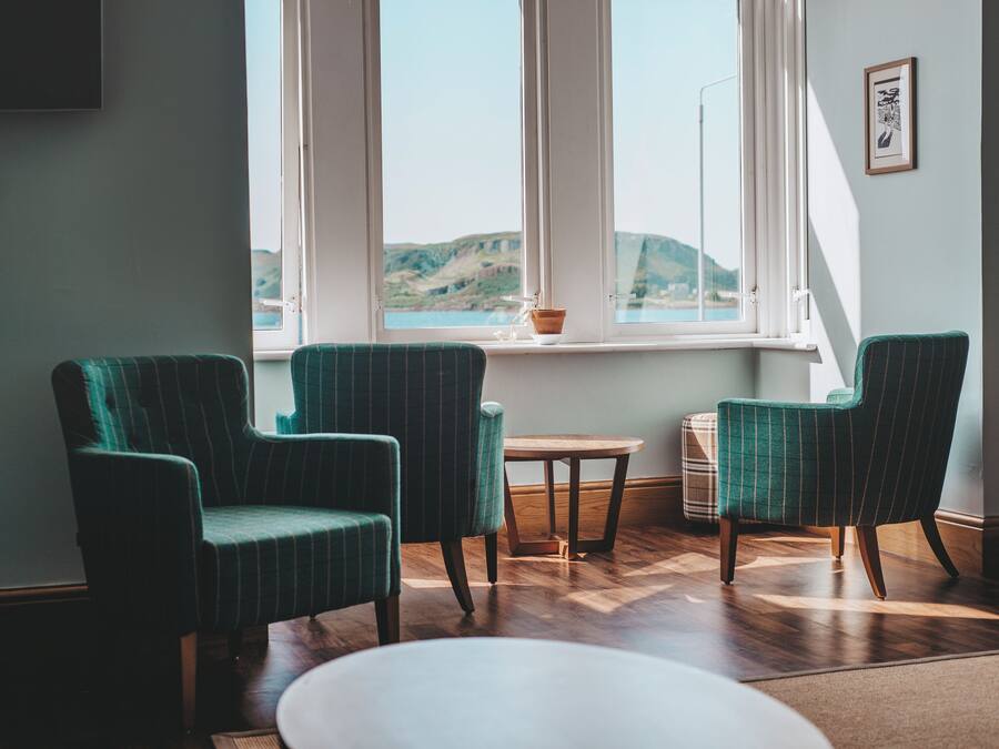 A room with two green upholstered chairs, a small round table, and a view of a coastal landscape through a window.