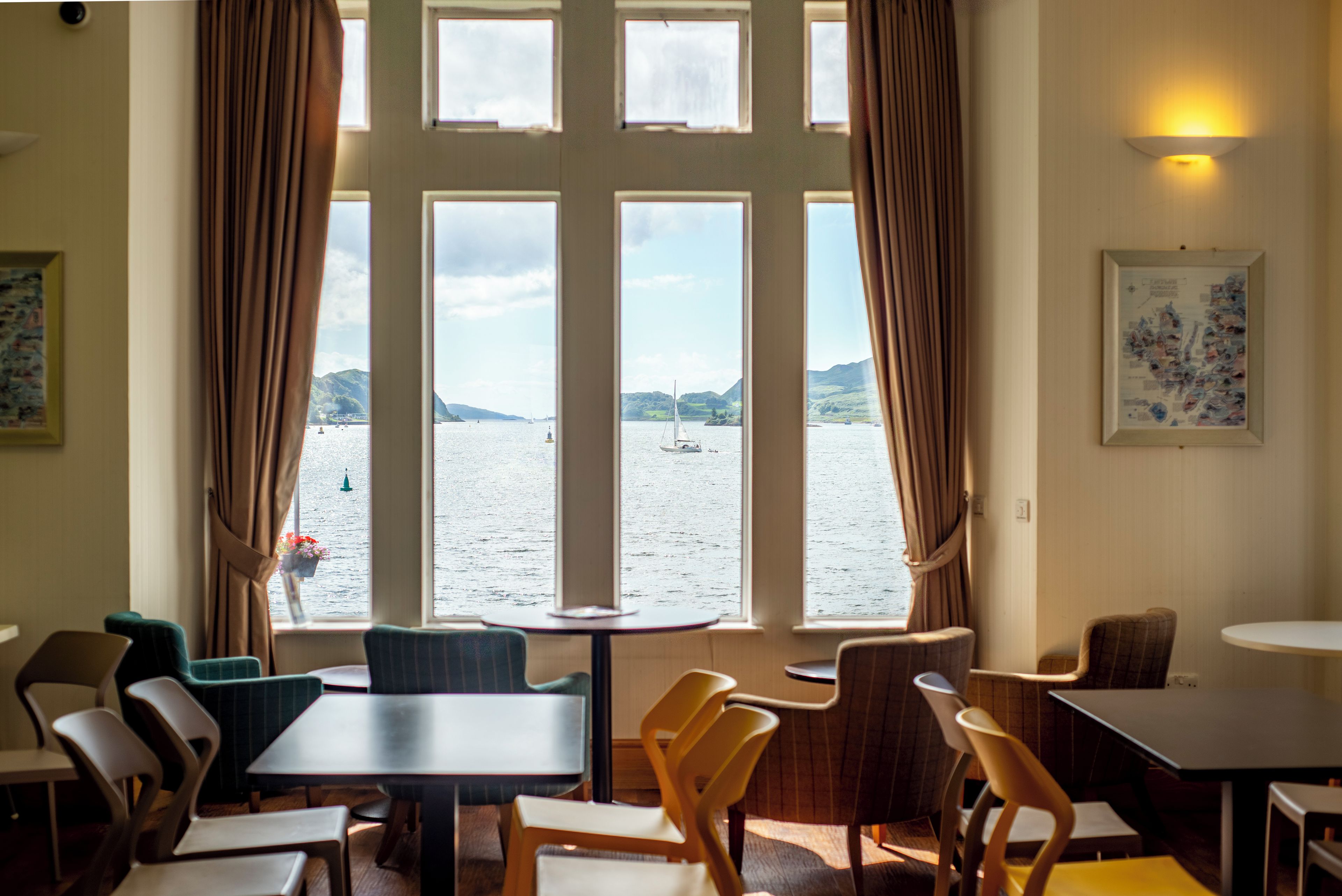A room with a large window overlooking a body of water, several tables and chairs arranged for dining, and a wall-mounted light fixture.