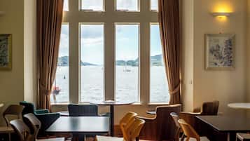A room with a large window overlooking a body of water, several tables and chairs arranged for dining, and a wall-mounted light fixture.