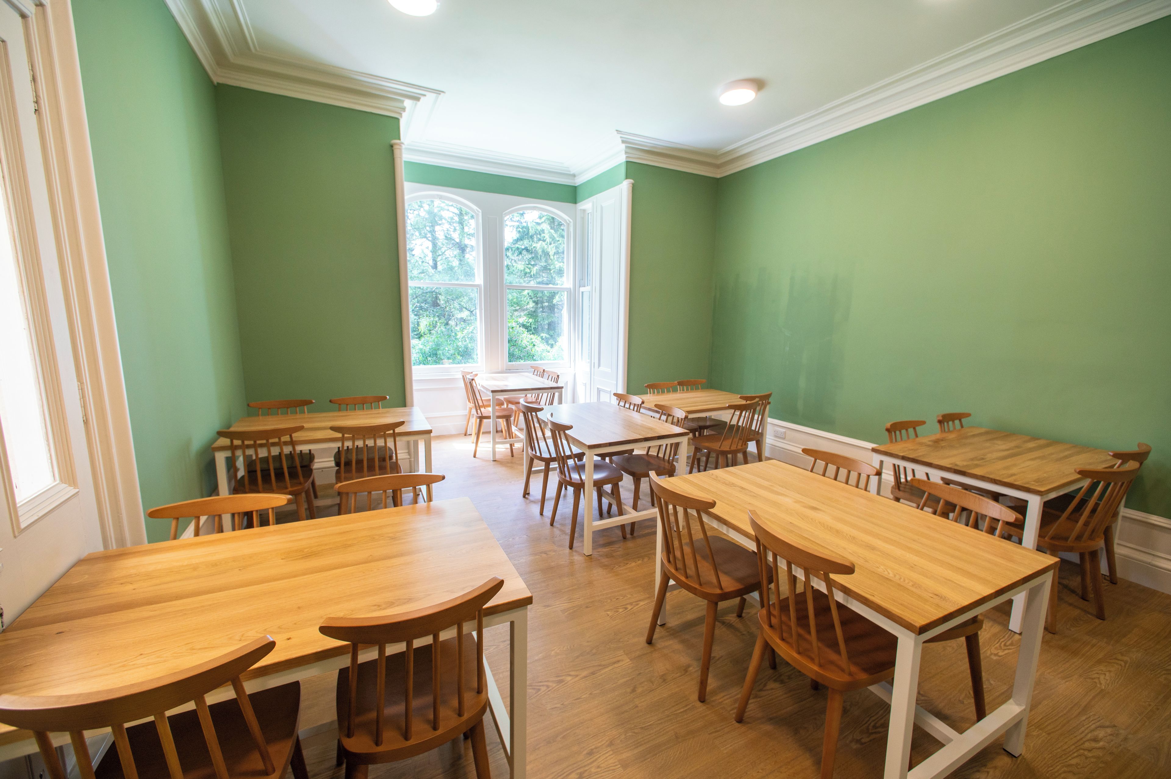 A dining area with multiple wooden tables and chairs, large windows with a view of trees, and a green wall.