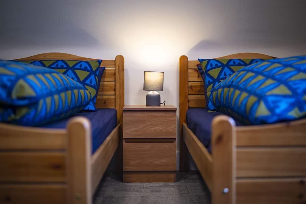 A small, cozy room with two wooden beds, each with a blue and yellow patterned pillow. A small wooden nightstand with a lamp is between the beds.