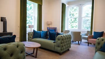 A living room with a green sofa, a round coffee table, and two armchairs. There is a fireplace on the left and a window with curtains on the right.