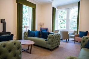 A living room with a green sofa, a round coffee table, and two armchairs. There is a fireplace on the left and a window with curtains on the right. - Braemar Youth Hostel (Ballater)