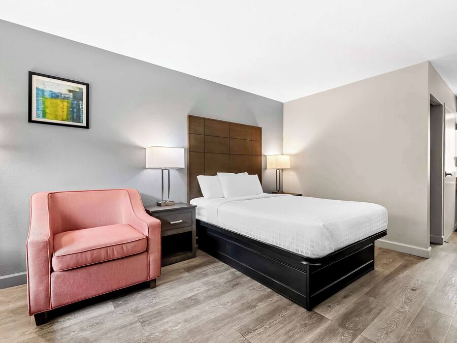 Une chambre d’hôtel moderne, avec un lit, un fauteuil rose, des tables de chevet avec des lampes et une œuvre d’art encadrée au mur.