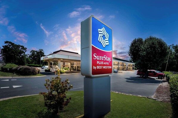 A SureStay PLUS Hotel sign with a modern design, located in front of a hotel building with a well-maintained lawn and parked cars.