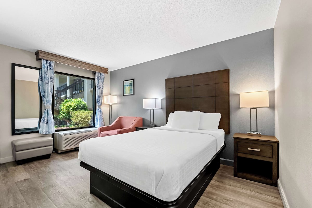 Ein Hotelzimmer mit einem großen Bett, einem Schreibtisch, einem Sessel, einem Fenster mit Vorhängen und Blick ins Grüne.