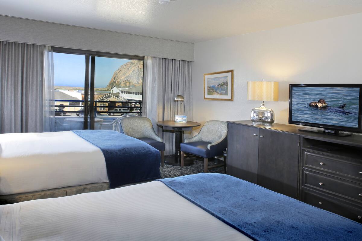 Quarto de hotel com duas camas, uma escrivaninha, uma cadeira, televisão e vista para uma montanha e um corpo de água.