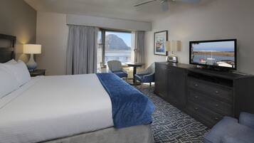 Ett hotellrum med en stor säng, ett skrivbord, en stol, en tv och utsikt över havet.