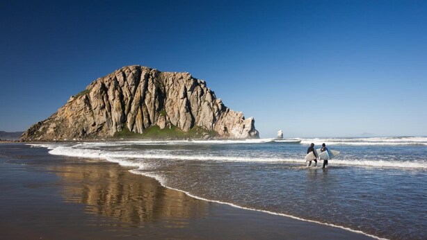 A rocky coastline with two people carrying surfboards, a clear sky, and calm waves. - Blue Sail Inn (Morro Bay)