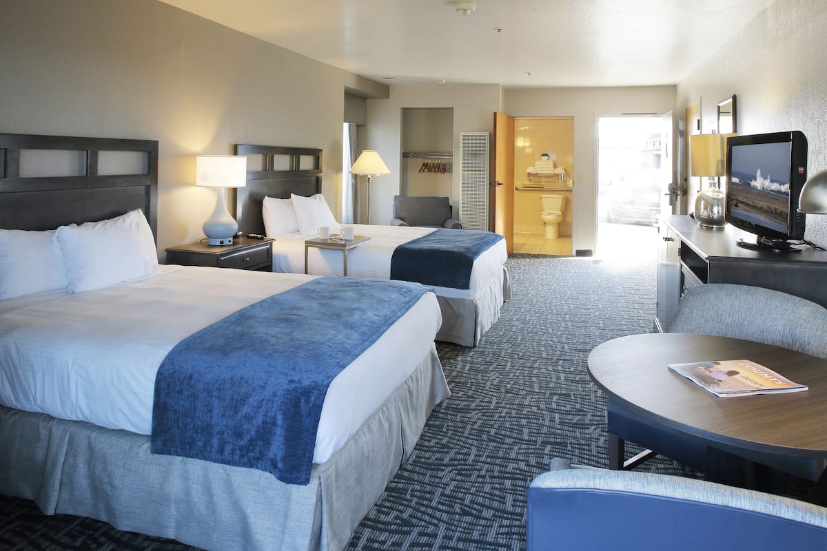 Quarto de hotel com duas camas, uma escrivaninha com computador, televisão e área de estar com uma revista.