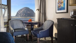 A hotel room with two blue armchairs, a small table with a lamp, a painting on the wall, and a view of a mountain and water through the window. - Blue Sail Inn (Morro Bay)