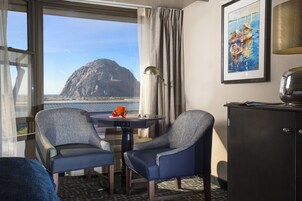 Habitación de hotel con dos sillones azules, una mesita con una lámpara, un cuadro en la pared y vistas a una montaña y al agua a través de la ventana.