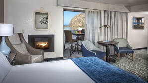 A hotel room with a bed, two armchairs, a small table, a fireplace, and a view of the sea. - Blue Sail Inn (Morro Bay)