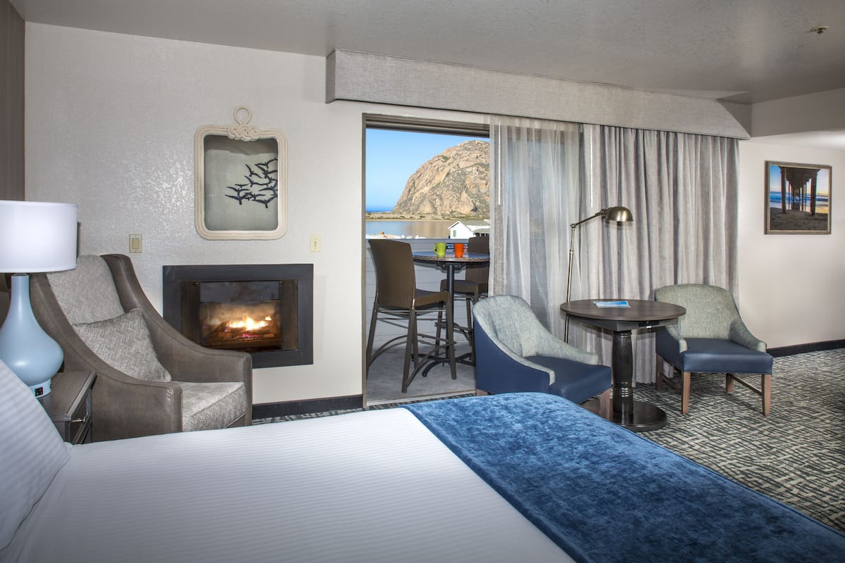 Quarto de hotel com uma cama, duas poltronas, uma mesinha, lareira e vista para o mar.