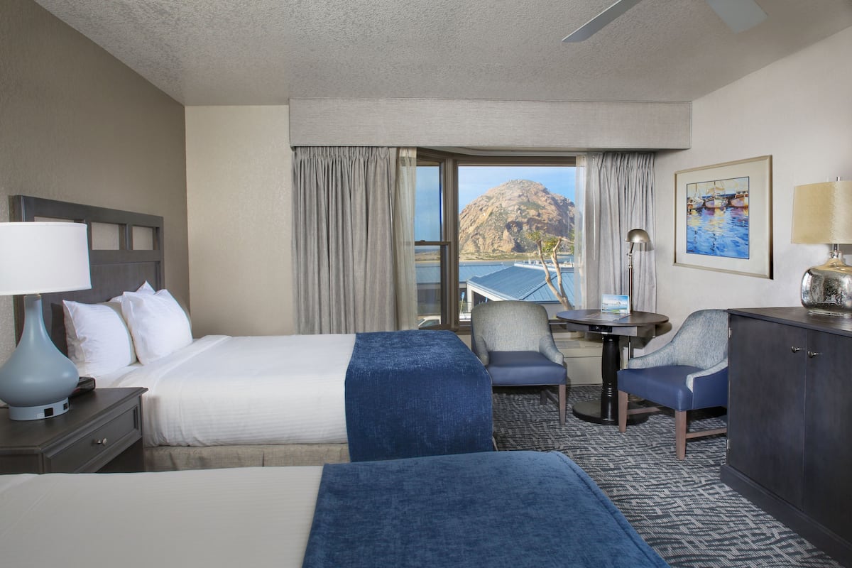 Quarto de hotel com cama, escrivaninha, duas cadeiras, abajur e vista para uma montanha e água.