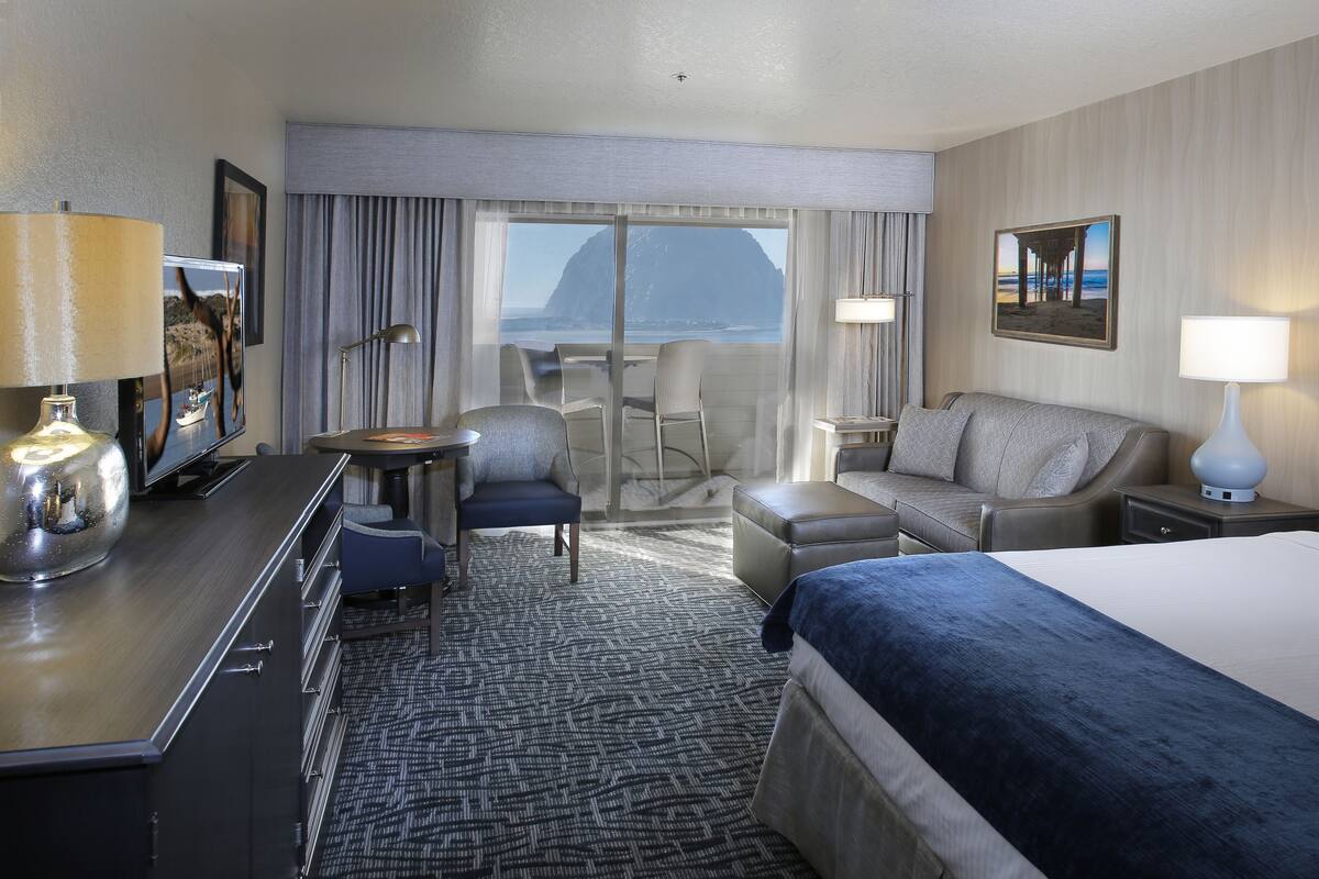 Quarto de hotel com cama, escrivaninha com luminária, cadeira, sofá e vista para o mar.