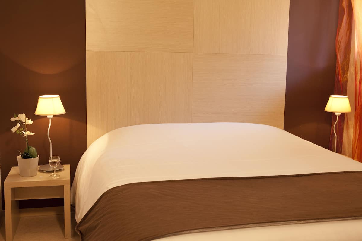 A neatly made bed with a brown headboard, a small bedside table with a lamp, and a potted plant.