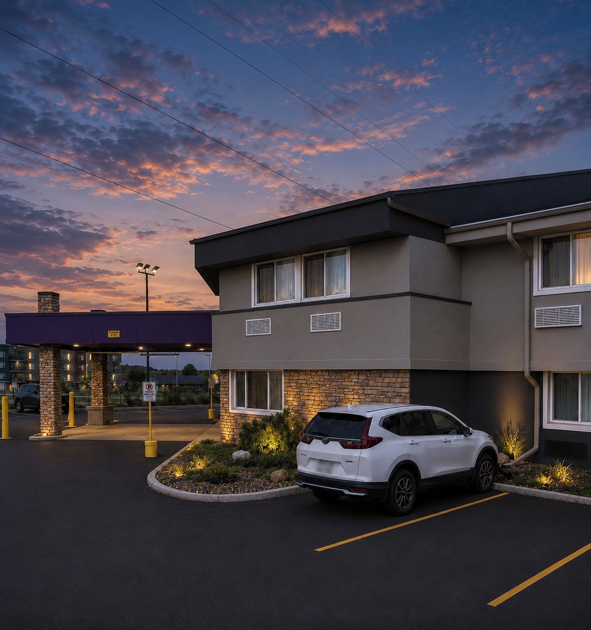 A modern hotel with a white SUV parked in the driveway, a no parking sign, and a sunset sky.